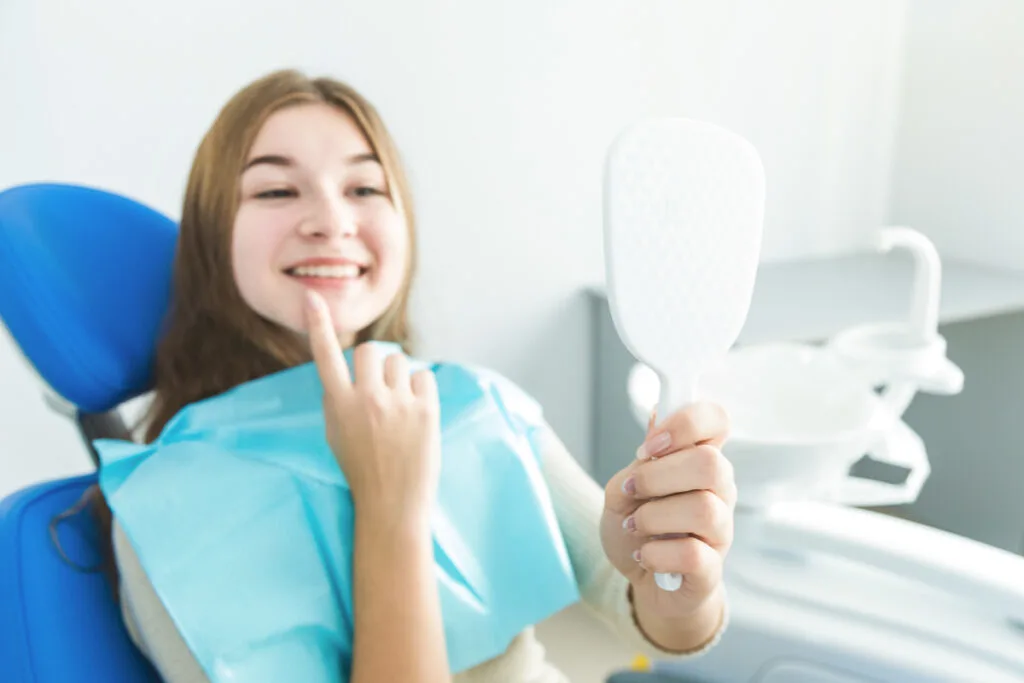 Frau hält beim Zahnarzt einen Spiegel in der Hand - schiefe Zähne bei Erwachsenen